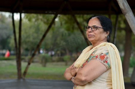 印度新德里摄影照片_在印度新德里的夏天，穿着米白色萨尔瓦卡米兹旁遮普西装的公园里，聪明自信的北印度高级女性站立、思考和双手交叉/折叠