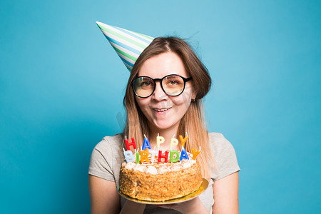 大生日蛋糕摄影照片_疯狂有趣的女孩戴着纸帽子和眼镜，在蓝色背景上拿着一个大生日蛋糕。