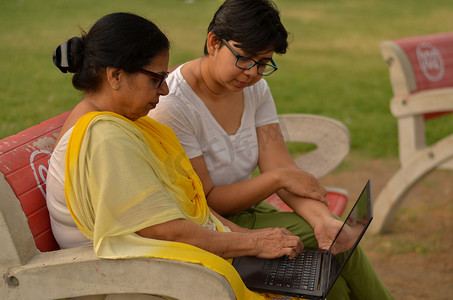 印度新德里摄影照片_在印度新德里的一个公园里，年轻的印度女孩在坐在红色长椅上的笔记本电脑上帮助一位印度老妇人的侧视图。