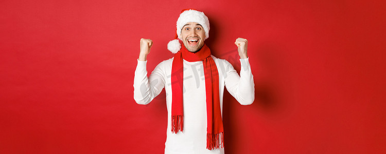 圣诞帽围巾摄影照片_戴着圣诞帽和围巾的快乐而兴奋的男人的画像，欢欣鼓舞并赢得了一些东西，庆祝新年，站在红色背景上