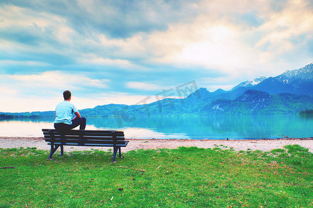 身穿蓝色衬衫的疲惫成年男子坐在山湖沿岸的旧木凳上