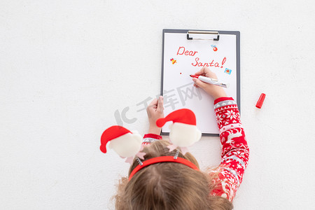 亲爱的圣诞老人信，圣诞卡。女孩穿着红色毛衣，拿着毡笔，在白色背景的白纸上写字。关于礼物的童年梦想。圣诞节和新年概念。寒假