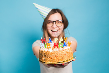 生日蛋糕海报摄影照片_特写滑稽模糊的积极女孩，戴着眼镜，戴着问候纸帽，手里拿着一个生日快乐蛋糕，站在蓝色背景上。