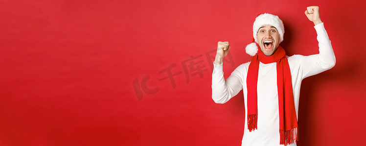 圣诞帽围巾摄影照片_戴着圣诞帽和围巾的快乐白人男子的形象，欢呼雀跃，举起双手，庆祝胜利或胜利，战胜红色背景