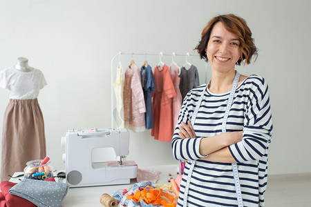 一位积极的年轻女裁缝师站在她的桌面背景上，带着配件和缝纫机的肖像。