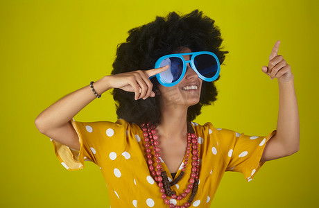美丽而微笑的女人，留着卷曲的非洲发型，戴着心形太阳镜，用黄色背景上的手指向右看和指示方向
