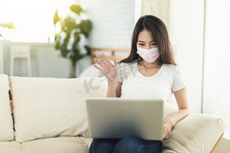 在家工作的年轻女性戴着口罩保护自己，坐在沙发上使用笔记本电脑进行远程办公，并与同事视频聊天