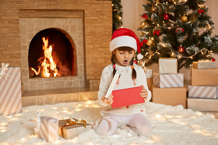 小女孩在圣诞节打开礼物时兴奋不已，女孩戴着圣诞老人的帽子坐在地板上，张着嘴，惊讶和惊讶，在客厅里摆出圣诞装饰的姿势。