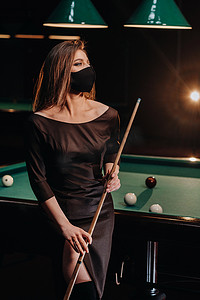 黑衣蒙面人摄影照片_台球俱乐部里的蒙面女孩手里拿着球杆