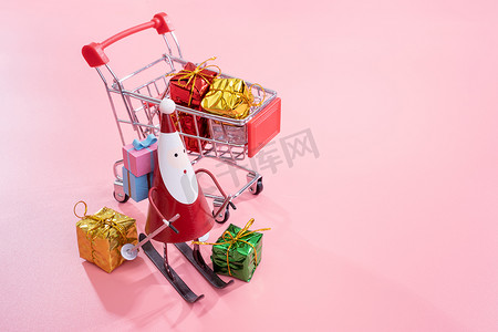 圣诞购物概念、带圣诞老人玩具的迷你红色购物车手推车和淡粉色背景中突显的礼盒、空白复制空间、特写