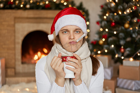 心烦意乱的女性戴着圣诞帽、围巾和毛衣，测量温度和喝热茶，用两只手拿着杯子，坐在壁炉和圣诞树附近。