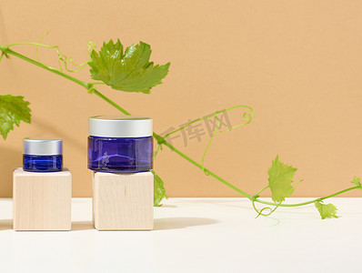 蓝色玻璃罐中的化妆品，灰色盖子放在由立方体制成的木制讲台上，后面是一枝绿叶葡萄。