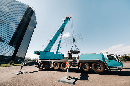 配重由一名面目全非的工人安装在一台大型蓝色汽车起重机上，准备在一座大型现代化建筑旁边的工地上工作。