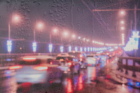 街道夜摄影照片_在被雨水覆盖的窗户后面的夜城里，移动的汽车和灯笼的模糊灯光反射在潮湿的沥青上。