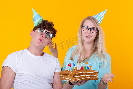 有趣的年轻夫妇戴着纸帽，拿着蛋糕，站在黄色背景下，做着傻脸，祝生日快乐。