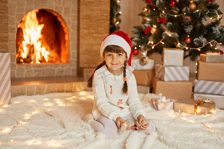 孩子在创作摄影照片_戴着圣诞老人帽和白色毛衣的迷人孩子坐在圣诞树附近的地毯上，手里拿着小玩具在壁炉上，看起来对着镜头微笑，在家里庆祝假期。