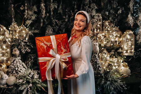 一个穿着白色连衣裙的女孩，手里拿着一件大圣诞礼物，背景是童话故事。在圣诞树和小房子的背景下，穿着白色衣服的微笑女人
