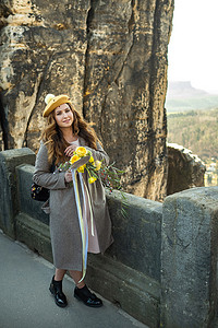 灰色外套摄影照片_在瑞士萨克森、德国、巴斯泰的山脉和峡谷背景中，一个身穿灰色外套和帽子、戴着一束鲜花的女孩