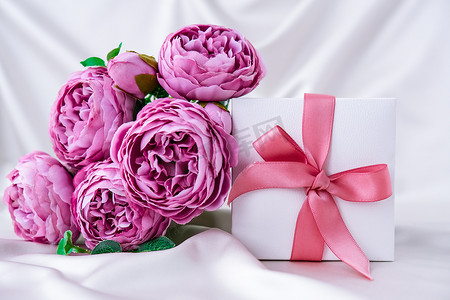 白色礼物盒，白色丝绸背景上有粉色丝带和紫色牡丹花。