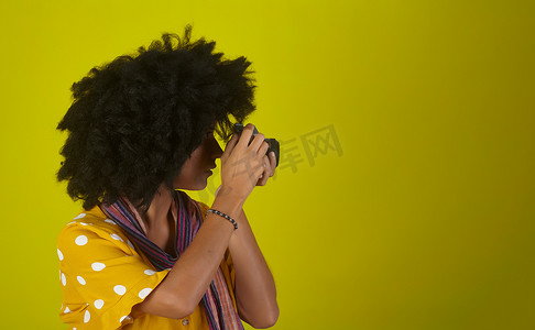 黄色背景中留着卷发爆炸头的美女，同时用复古胶片相机拍照