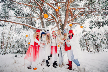 冬天水果摄影照片_一大群带着橘子的女孩站在冬天的森林里。在白雪覆盖的森林里，穿着红白相间水果的女孩