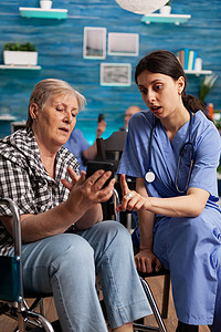 护士帮助坐在轮椅上的退休老年妇女在社会服务期间使用智能手机