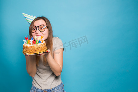 特写有趣的积极女孩，戴着眼镜，戴着问候纸帽，手里拿着一个生日快乐蛋糕，站在蓝色背景上。