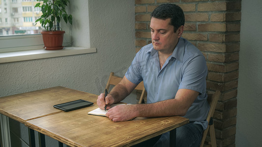 成年男性坐在咖啡馆里，拿着日记和平板电脑，在笔记本上记笔记。