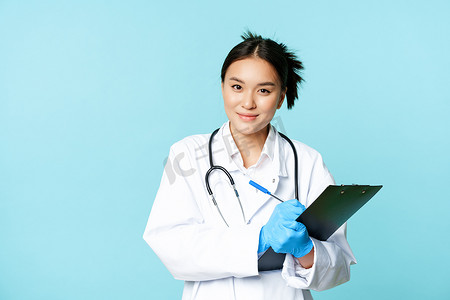微笑的亚洲医务工作者，女医生写下病人信息，拿着笔和剪贴板，站在蓝色背景上的制服