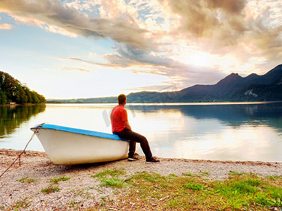 身穿红衬衫的疲惫男子坐在山湖沿岸的旧渔船上。