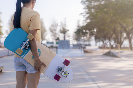 滑板坡道摄影照片_关闭年轻女性手持滑板，在公共滑板坡道公园背景上冲浪滑冰。