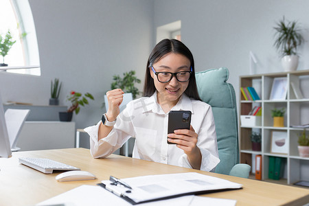 年轻的亚洲女商人坐在办公室的桌面上阅读电话后取得了成就。