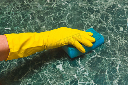 家庭主妇的手戴着黄色家用手套，正在厨房里用海绵摩擦绿色大理石表面