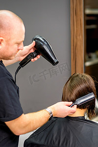 男性美发师在美发沙龙用黑色吹风机和黑色圆刷吹干年轻白人黑发女性的短发。