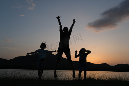 日落时小女儿和母亲在湖边跳跃的剪影。