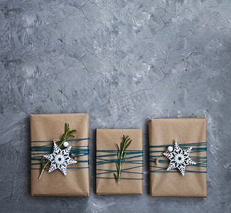 三件用牛皮纸包裹的圣诞礼物，用蓝色线包裹，灰色混凝土背景上有白色雪花和迷迭香枝条。