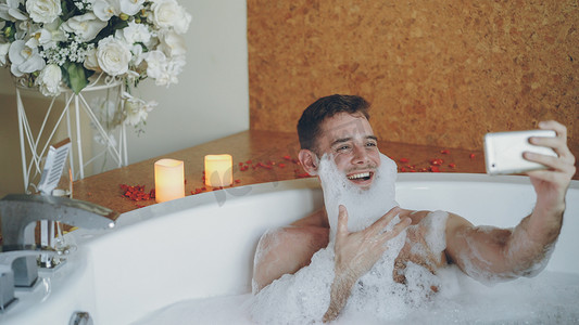 胡子上有泡沫的快乐男人正在现代水疗沙龙的热水浴缸里用智能手机自拍。