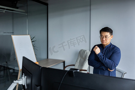 认知的人摄影照片_严肃而有思想的亚洲工程师看着两台显示器，办公室工作人员的特写照片，在现代办公楼内工作的男人，戴着眼镜和休闲衬衫的商人