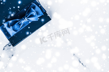 蝴蝶结蓝色礼盒摄影照片_冬季节日礼盒，配有蓝色丝绸蝴蝶结、大理石背景上的雪花，作为豪华美容品牌的圣诞和新年礼物，平铺设计