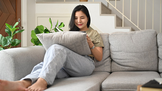 穿着休闲服的轻松年轻女性在沙发上使用数字平板电脑，在家度过闲暇时光