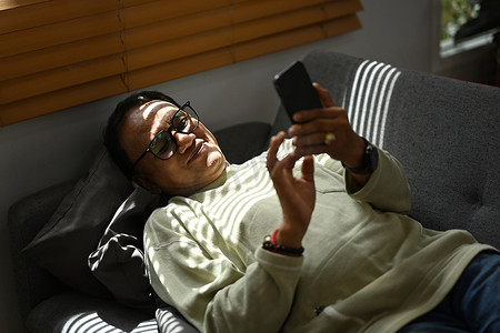 机上老人摄影照片_快乐的老人在家沙发上休息时阅读消息或在智能手机上查看社交媒体