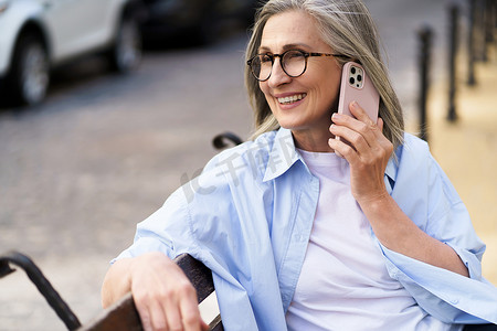 享受夏季时光，头发灰白的成熟女性拿着智能手机坐在欧洲老城街道的长凳上打电话。