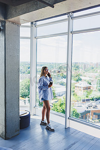 现代女性自由职业者站在大窗户前喝咖啡，打电话。