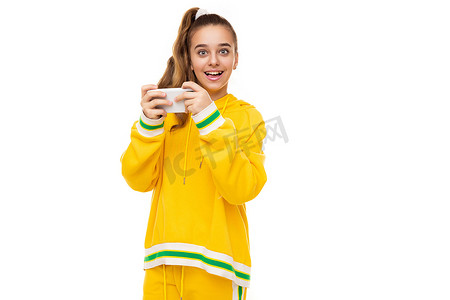 手机外壳摄影照片_照片中，一个美丽微笑的快乐女孩，黑发，马尾辫，穿着时尚的黄色运动服，绿色条纹，正在玩手机，手机的白色外壳隔离在白色背景上，有可用的文本空间