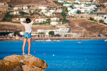 壮丽摄影照片_悬崖边上年轻快乐的女孩，可以欣赏希腊米科诺斯岛古老小村庄的壮丽景色