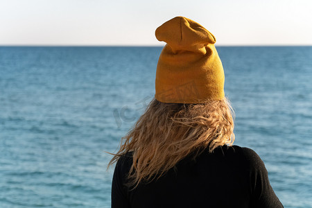 一个穿着黑色运动夹克和黄色帽子的金色卷发女孩正在看着大海。