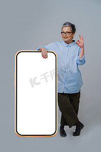 巨大手机摄影照片_戴眼镜的灰发成熟女商人靠在巨大的智能手机上，智能手机上有白色屏幕，穿着白色背景中突显的超大蓝色衬衫。