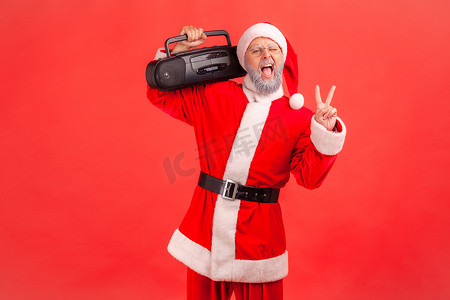 积极有趣的​​老人，留着灰色的胡子，穿着圣诞老人的服装，站着，拿着显示 v 形手势的录音机，庆祝圣诞晚会。