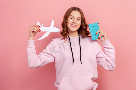 纸飞机粉色摄影照片_身穿连帽衫的快乐卷发少女的画像，微笑着在镜头前展示护照和纸飞机，旅游