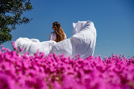 一位穿着白色飘逸长裙的漂亮女人住在一片美丽的田野附近，那里有粉红色的花朵后视